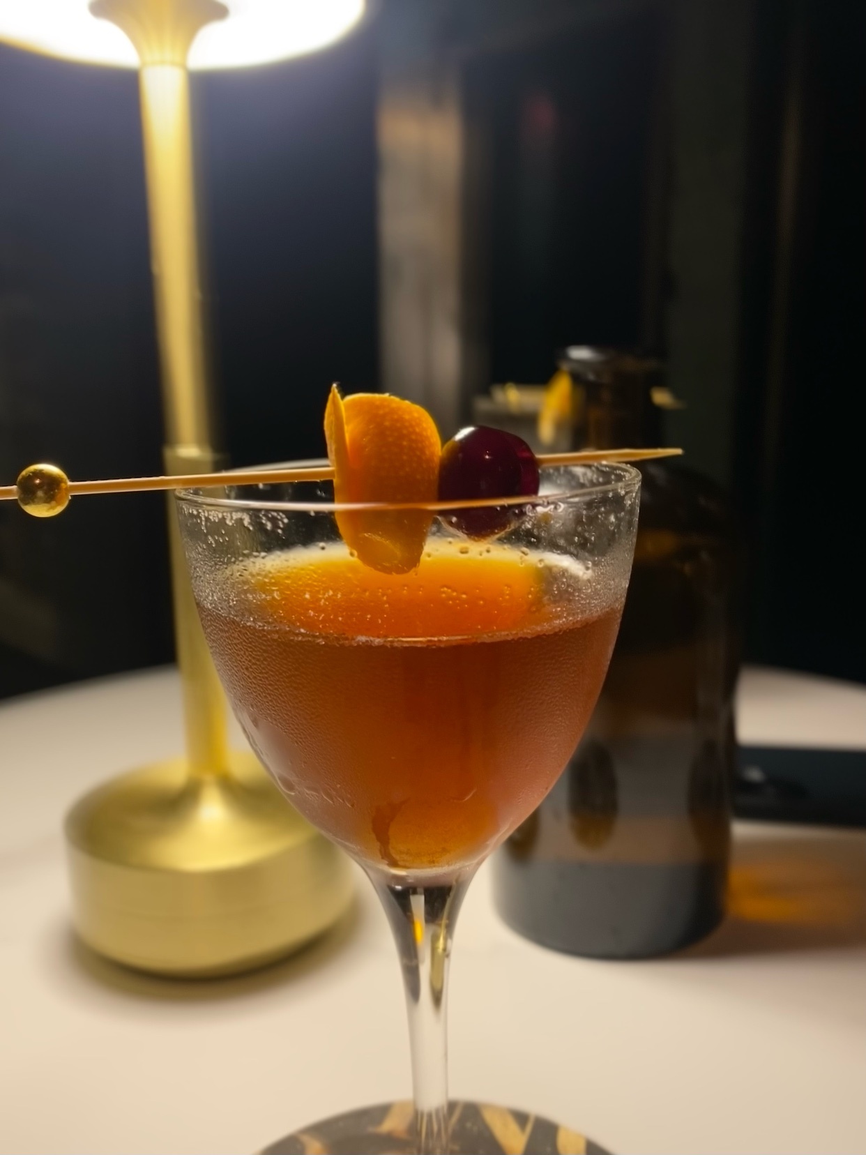 A sophisticated cocktail garnished with an orange peel and a cherry sits in a martini glass. The drink is amber in color and appears to be a classic Manhattan, with a bottle of beer blurred in the background.
