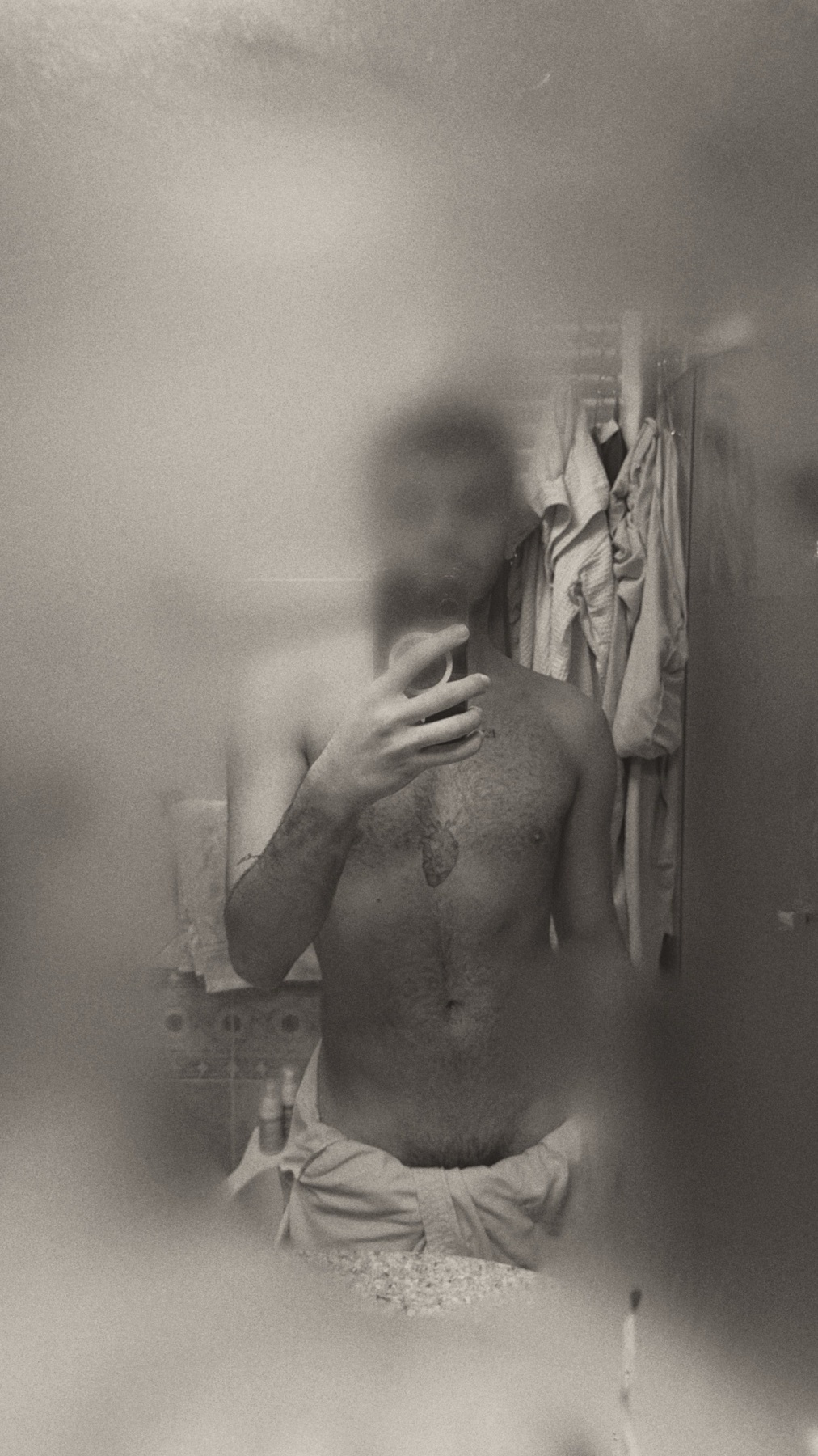 A man takes a mirror selfie, shirtless, in a bathroom. The image is in black and white.