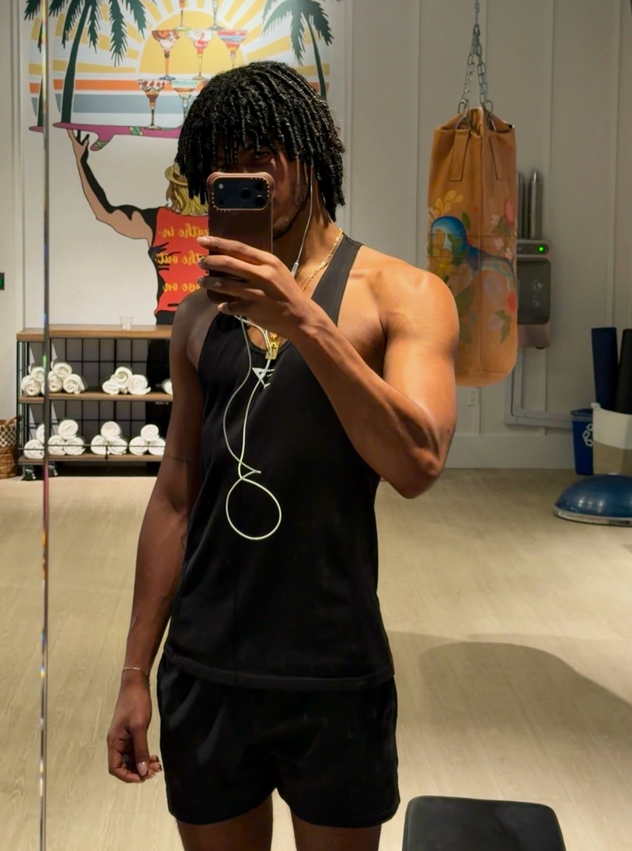 A man with dreadlocks takes a mirror selfie in a gym, wearing a black tank top and shorts. He is flexing his arm, showcasing his muscular physique.