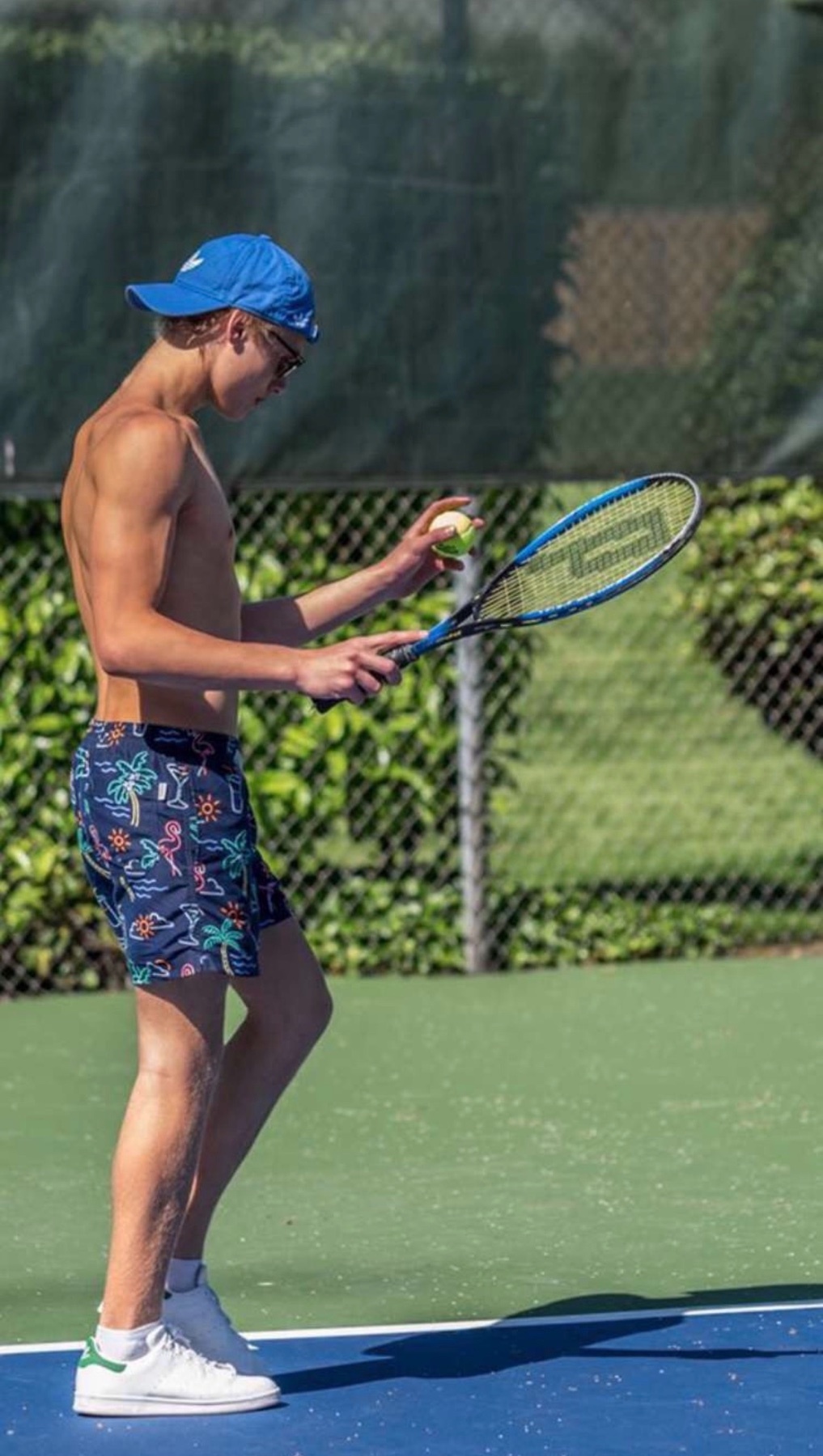 A shirtless young man in swim trunks and a blue hat stands on a tennis court, holding a tennis racket and ball. He appears to be preparing to serve.