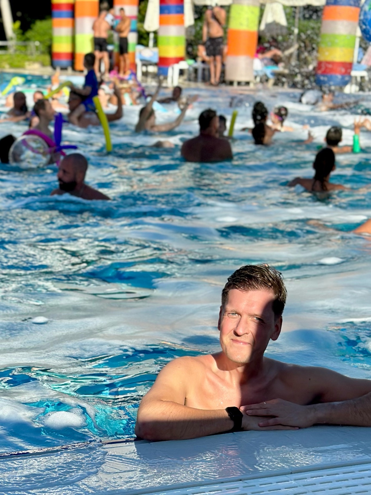 A man smiles at the camera while in a pool. Other people are in the pool, enjoying a pool party.