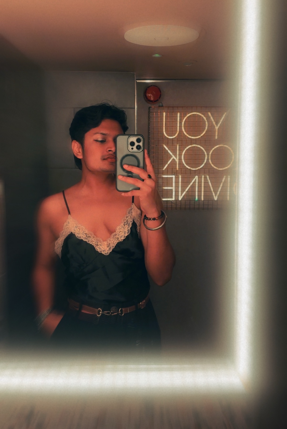 A person takes a mirror selfie in a dimly lit room, wearing a black satin camisole with lace trim and a belt. A neon sign in the background reads "YOU LOOK DIVINE".