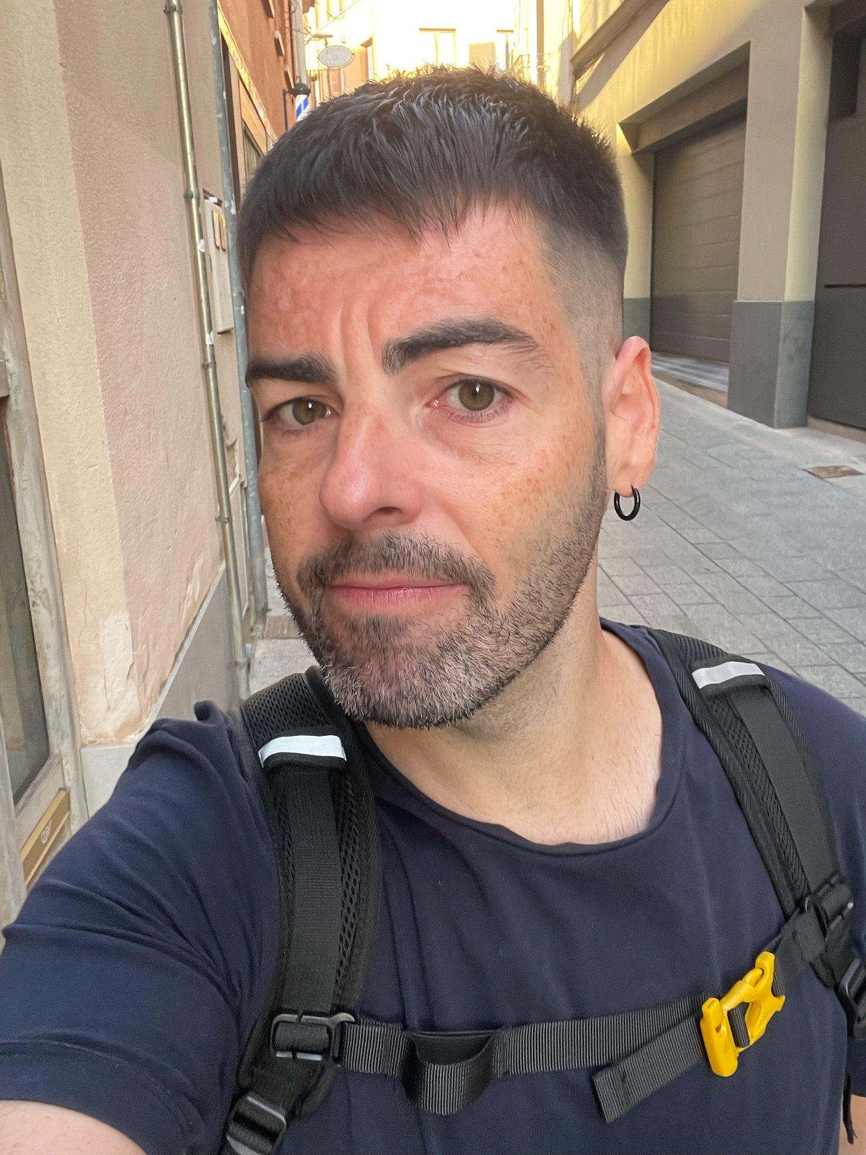 A man with short, dark hair and stubble poses for a selfie in a narrow street. He is wearing a dark t-shirt and a backpack with yellow accents.