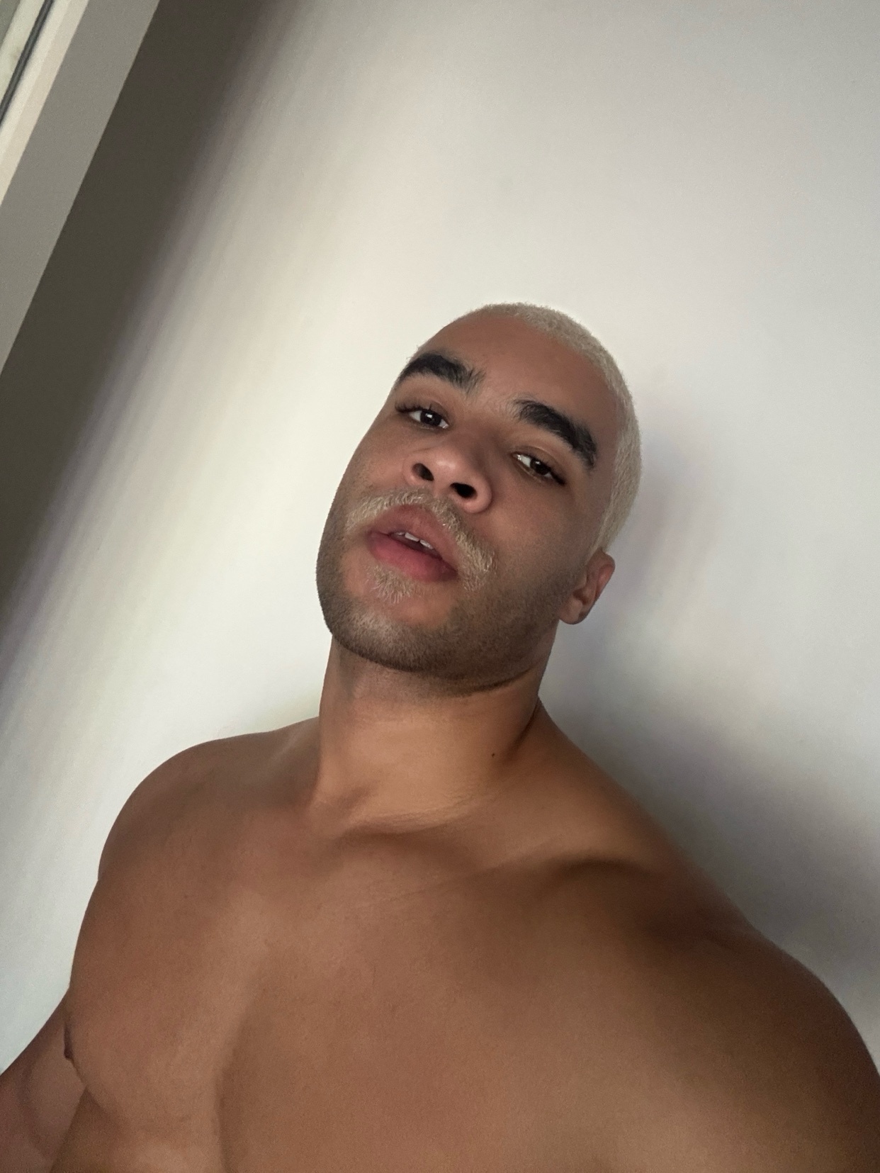 A shirtless man poses for a selfie, showcasing his muscular physique. He has blonde hair and a slight stubble.