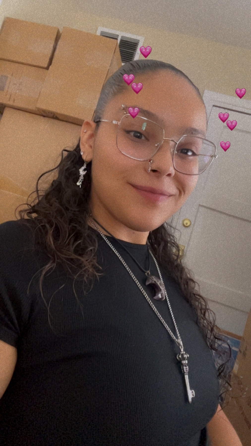 A young woman with curly hair and glasses smiles for a selfie. She is wearing a black shirt and several necklaces and earrings. Behind her are stacks of cardboard boxes.
