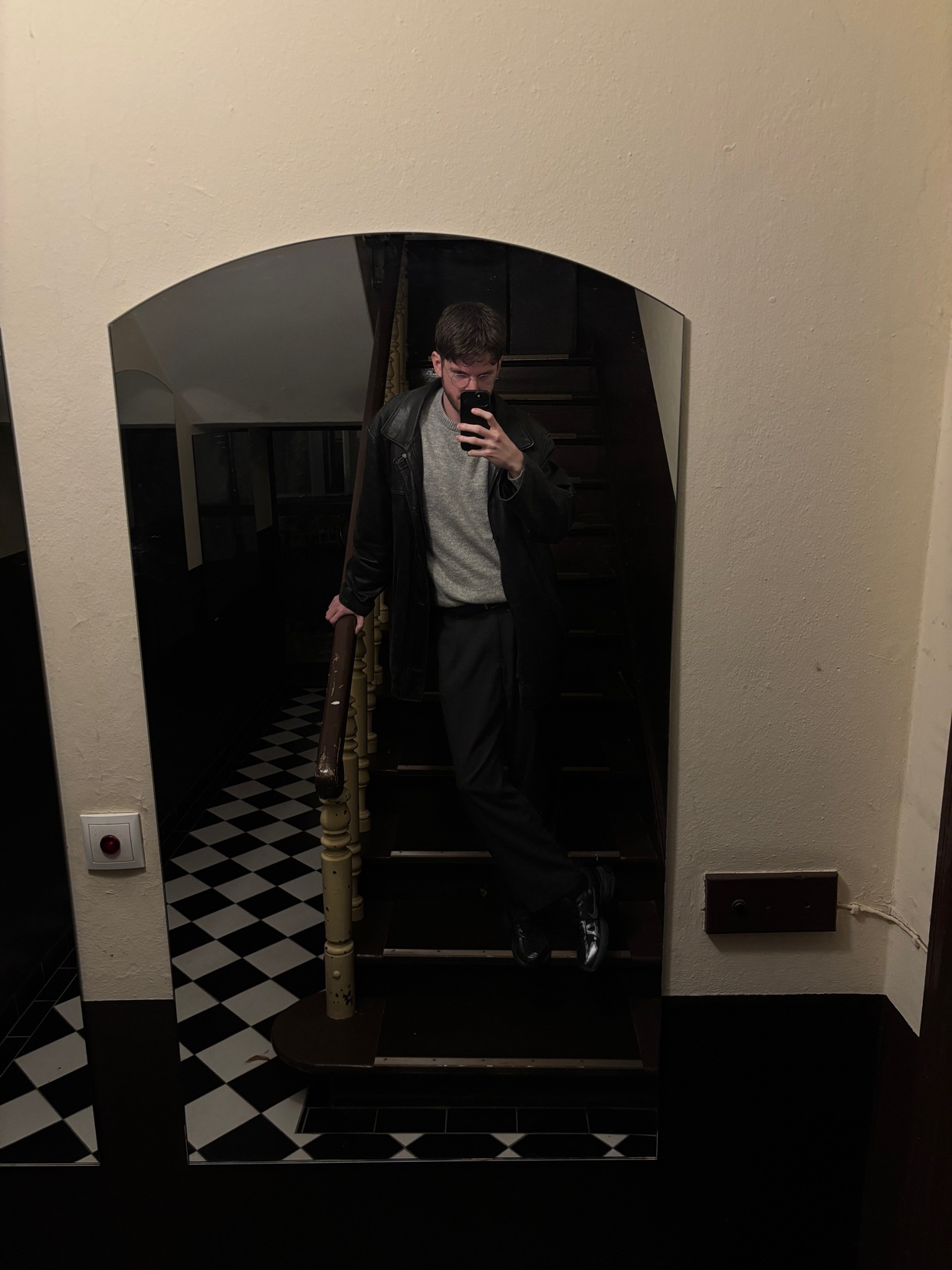 A man takes a mirror selfie on a staircase. He is wearing a leather jacket, grey sweater, and black pants.