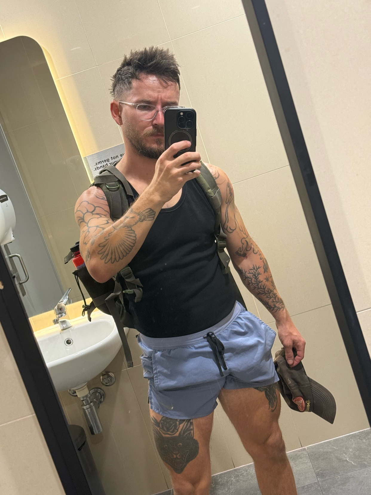 A man with tattoos and glasses takes a selfie in a bathroom, wearing a tank top, shorts, and a backpack.