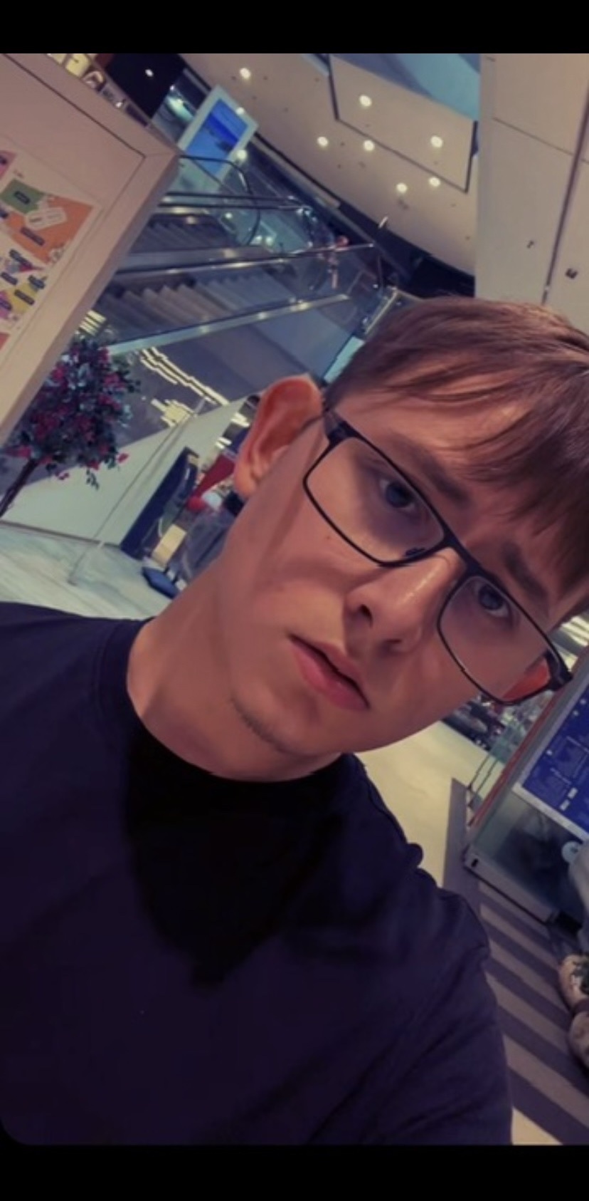A young man takes a selfie indoors. He is wearing glasses and looking at the camera.