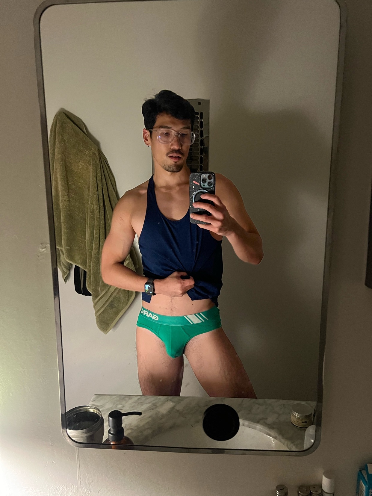 A man in a tank top and green underwear takes a mirror selfie. He is wearing glasses and has a mustache.