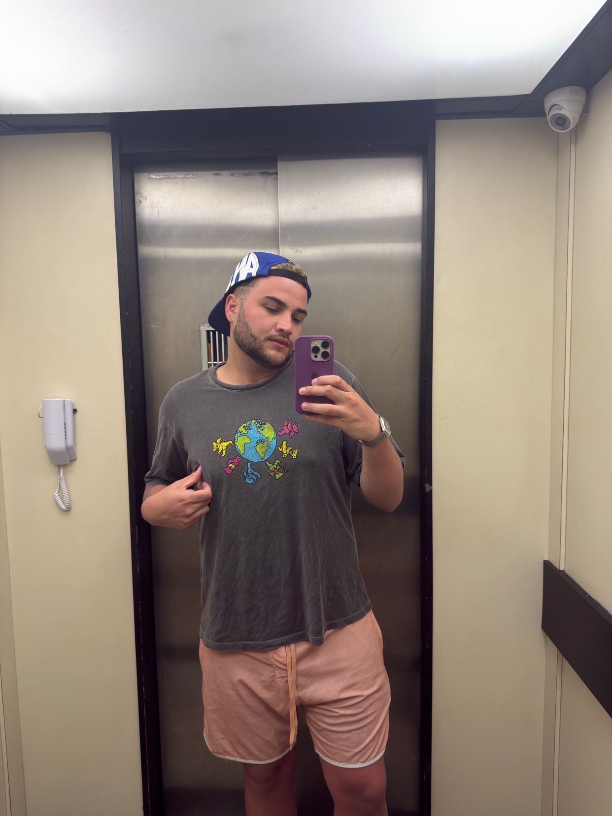 A man in a baseball cap, t-shirt, and shorts takes a selfie in an elevator.