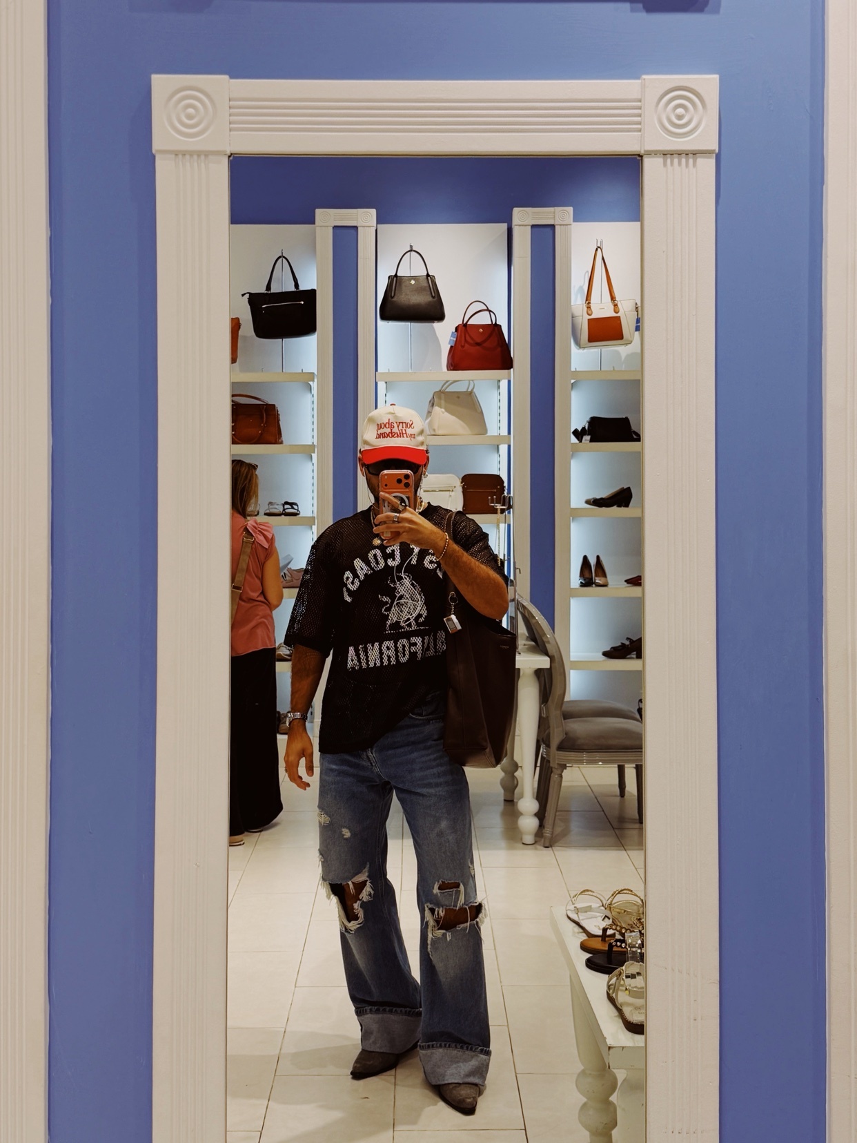 A man takes a mirror selfie while shopping. He is wearing a t-shirt, ripped jeans, and a cap.