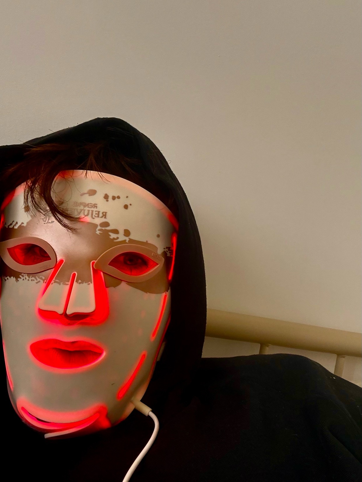 A person wearing a red LED light therapy mask and a black hoodie poses for a selfie against a plain wall.