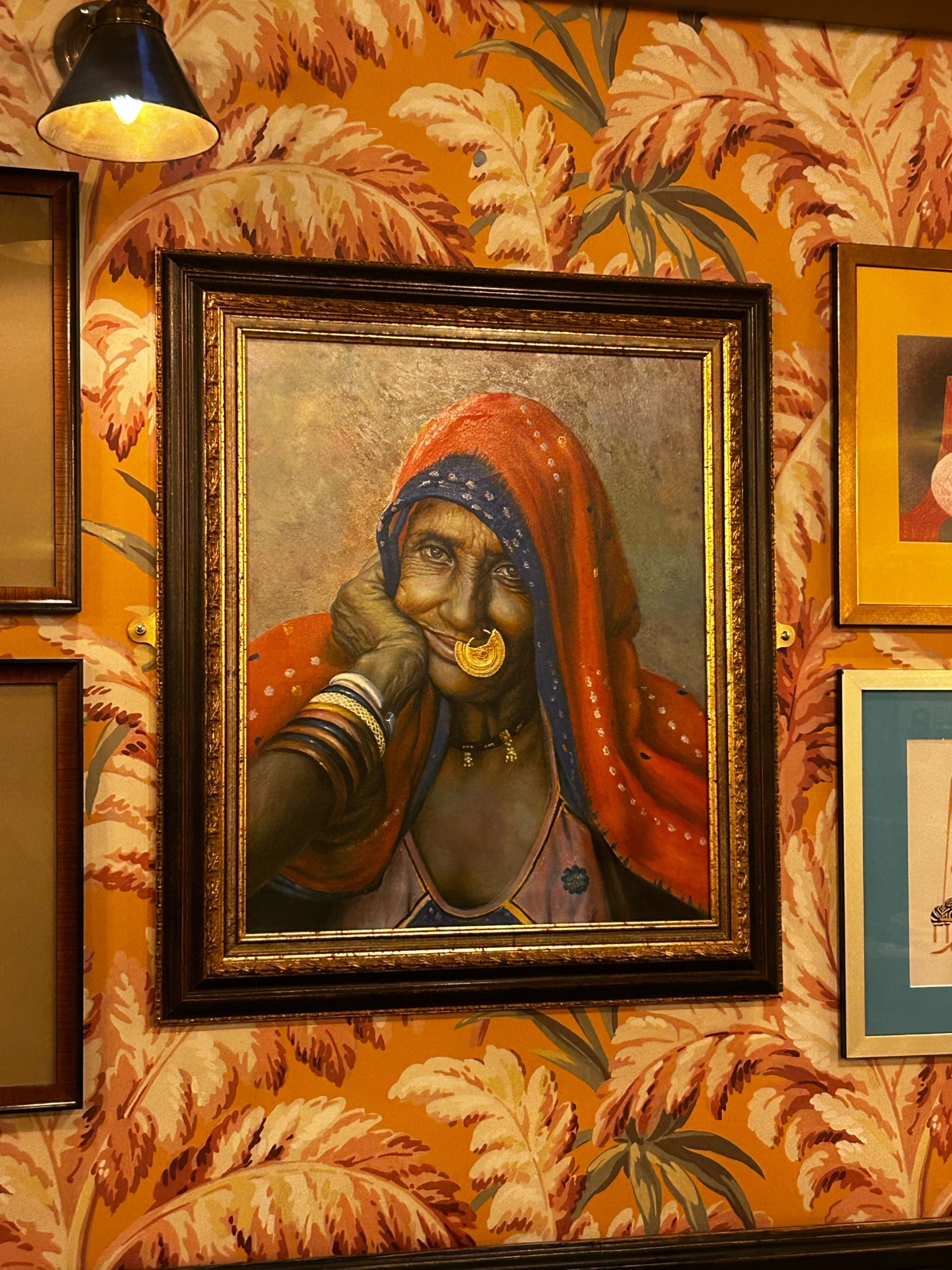 A framed painting of an Indian woman wearing a red headscarf and traditional jewelry is displayed on a wall with vibrant orange floral wallpaper.