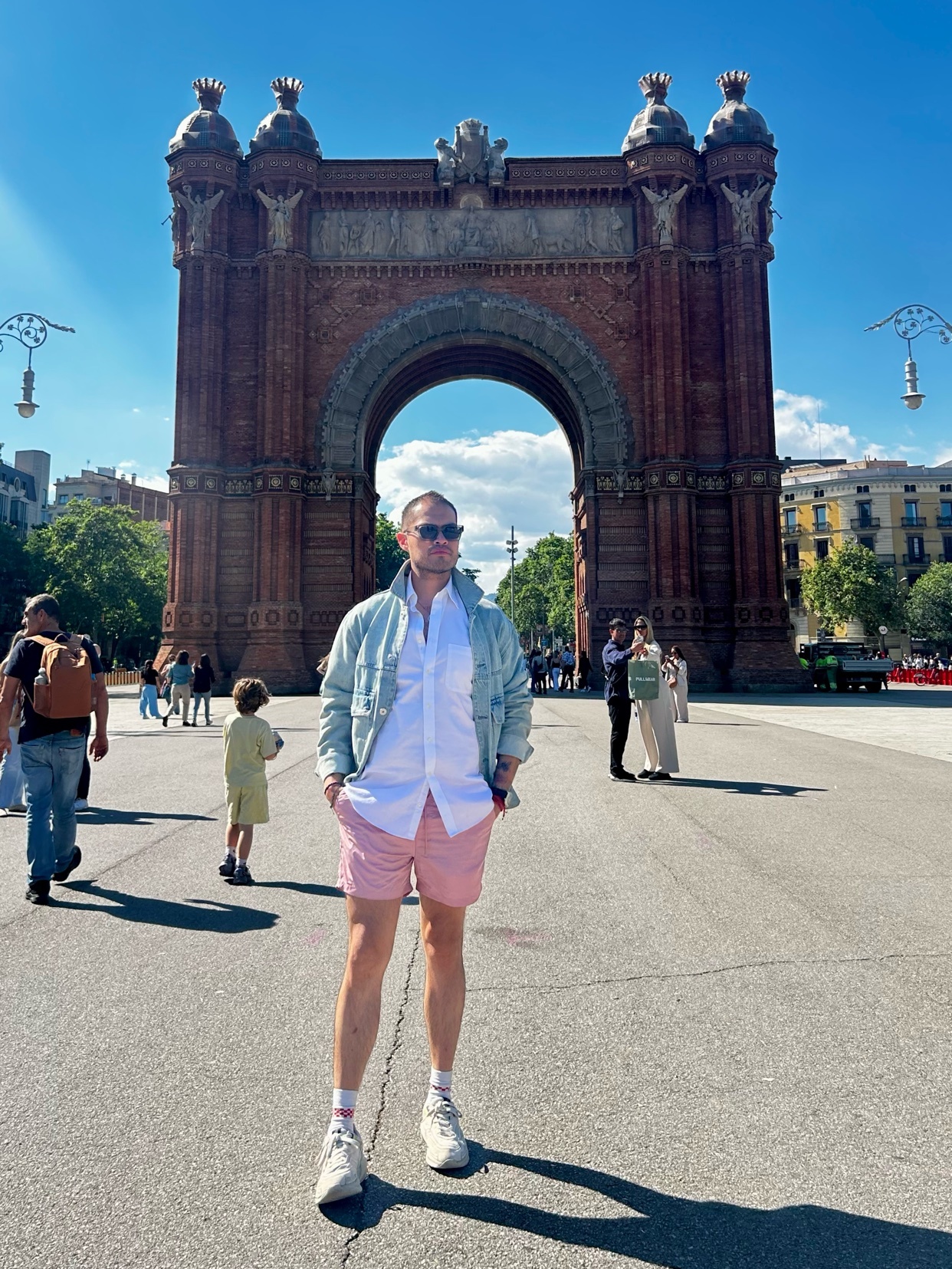 A man poses in front of the Arc de Triomf in Barcelona on a sunny day. He is wearing a white button-down shirt, pink shorts, and sunglasses.