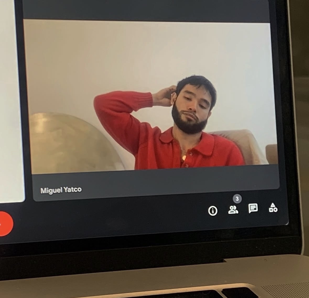 A man with a beard and dark hair is seen on a laptop screen during a video call. He is wearing a red sweater and has his arm behind his head.