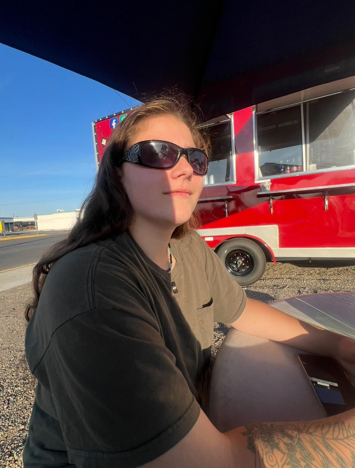A woman wearing sunglasses sits outside near a red food truck. The photo is taken during the day.