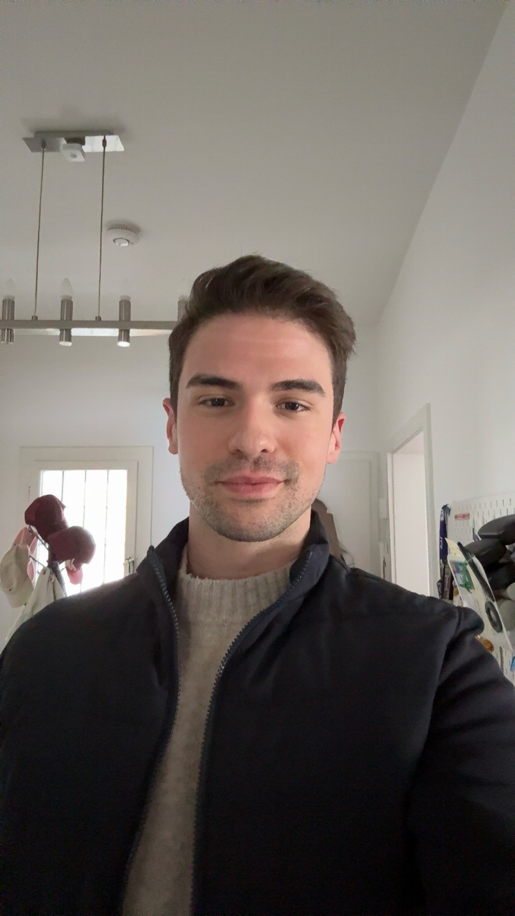 A man takes a selfie indoors. He is wearing a jacket and sweater.
