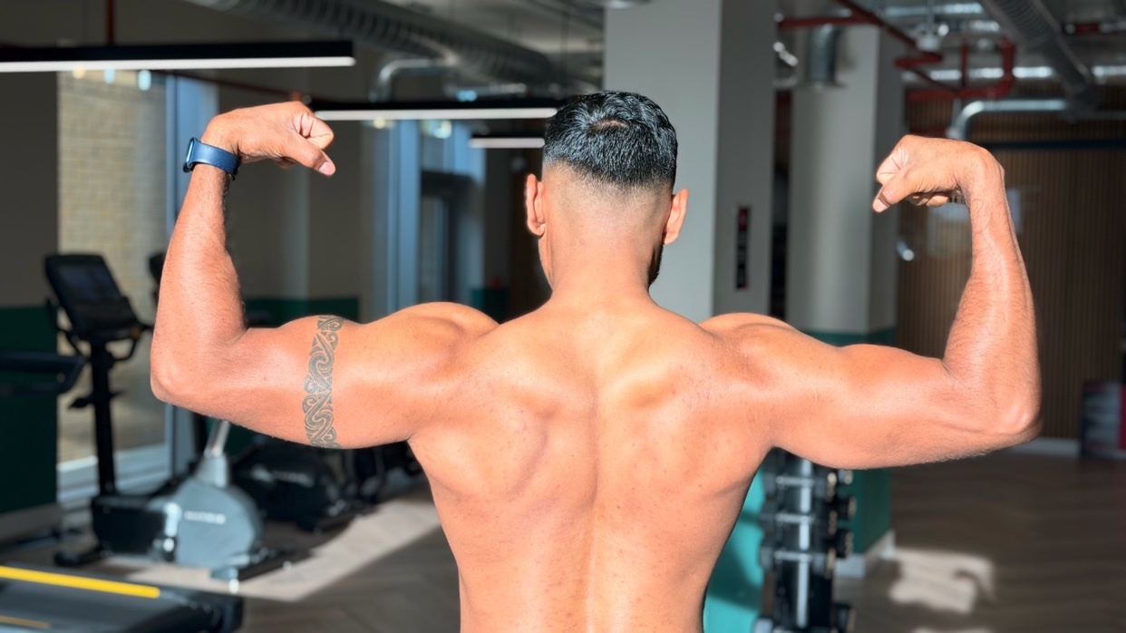 A man with a muscular back and biceps flexes his arms in a gym setting. He has a tattoo on his left arm.