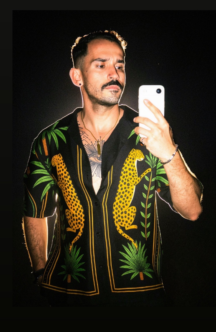A man takes a selfie in a black short-sleeved shirt featuring a bold leopard and palm tree print. He has tattoos visible on his chest and arms.