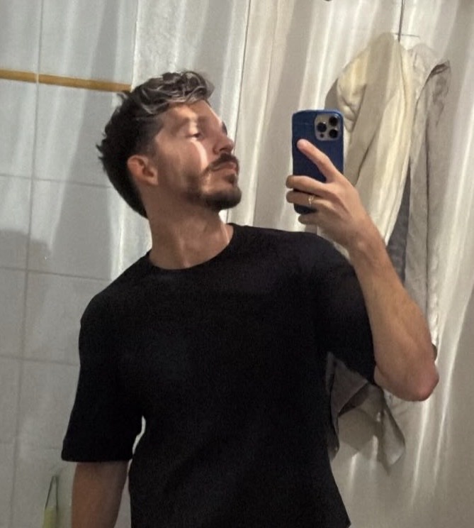 A man takes a mirror selfie. He is wearing a black t-shirt and has dark hair and a mustache.