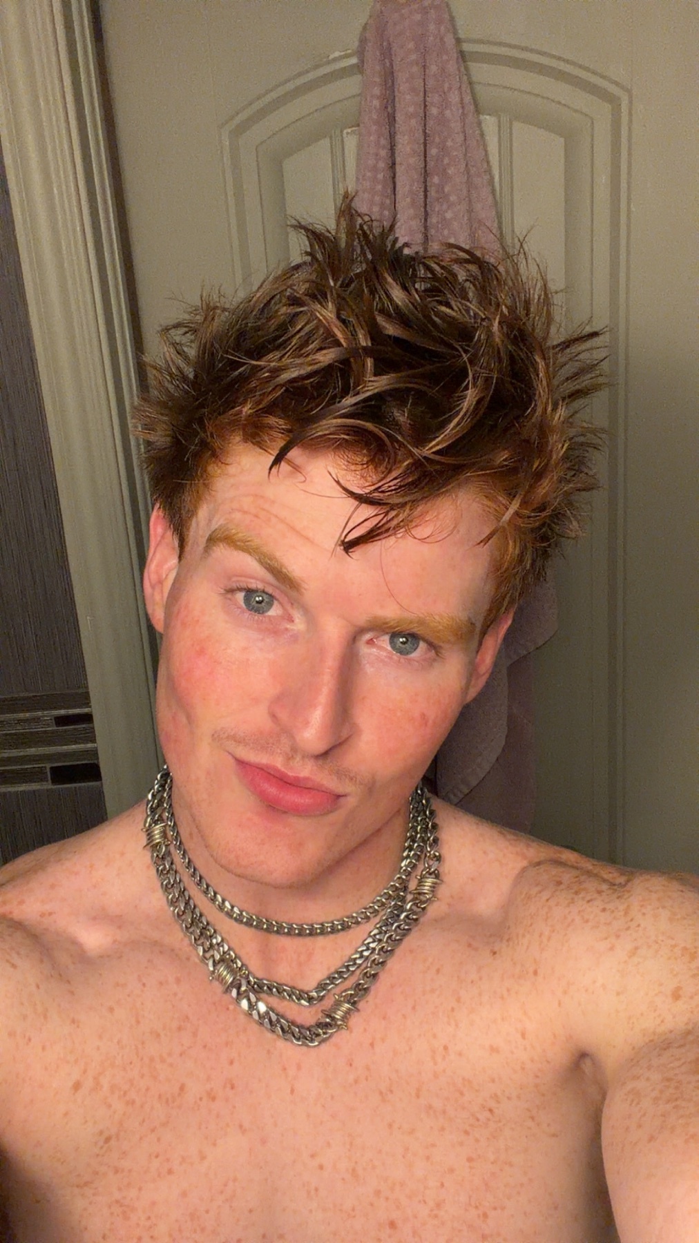A close-up selfie of a man with spiky red hair and freckles. He is wearing a multi-layered silver chain necklace.
