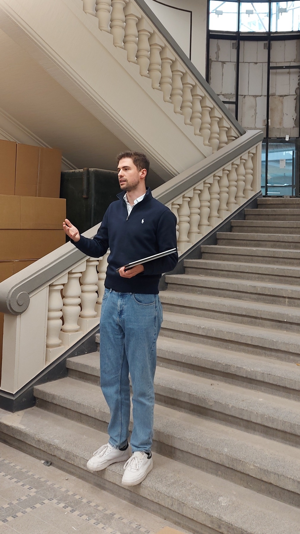 A man stands on a grand staircase, holding a laptop. He is dressed in casual business attire.