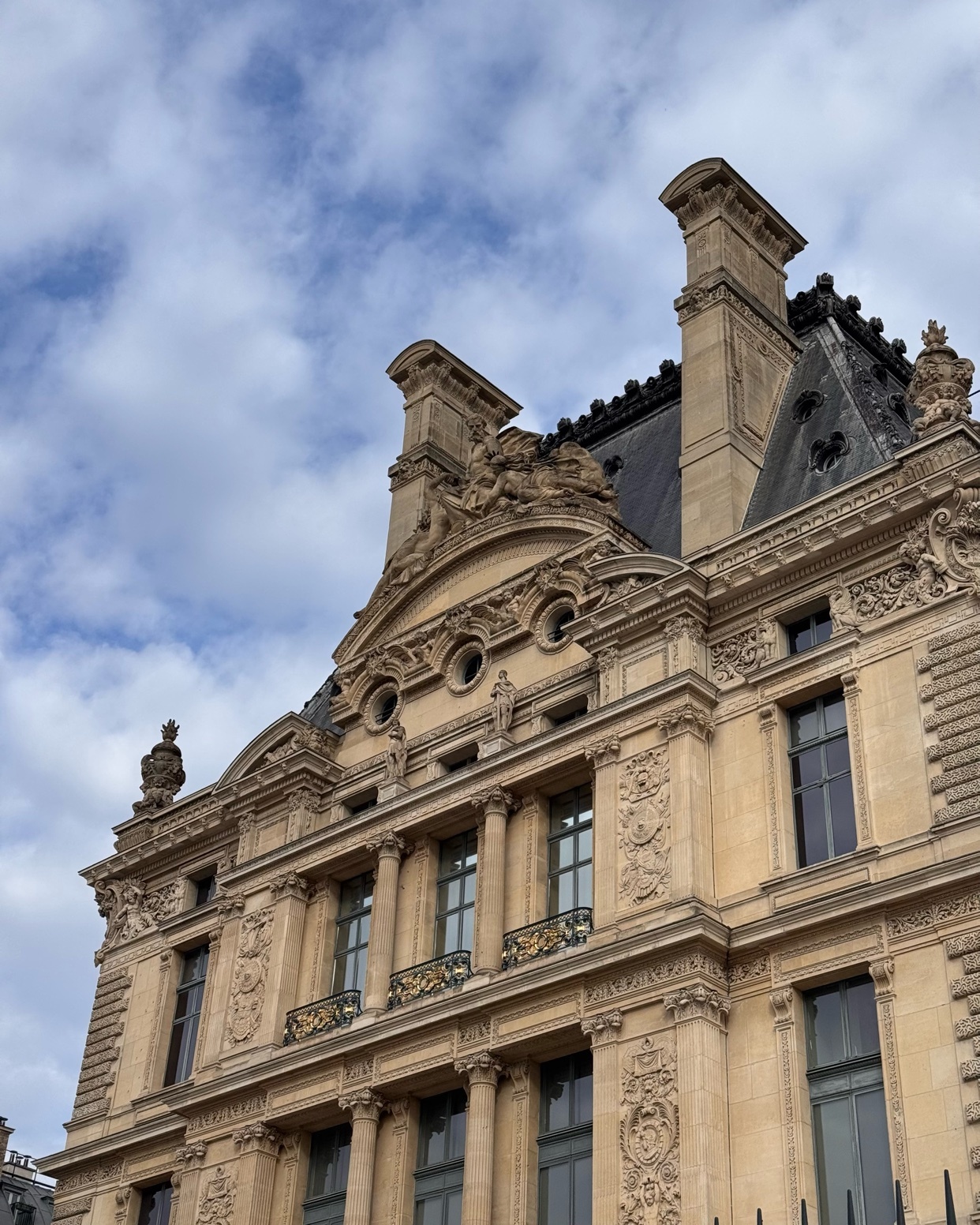 A close-up, low-angle shot of the ornate facade of a classical building, likely the Louvre Museum in Paris, under a cloudy sky. The intricate stone carvings and mansard roof showcase detailed French architecture.