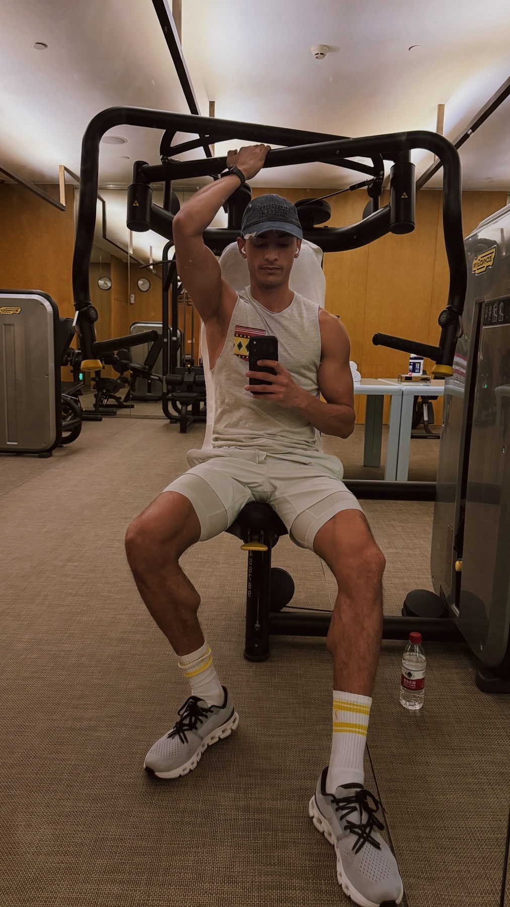 A man takes a mirror selfie at the gym while working out. He is wearing a tank top, shorts, and a cap.
