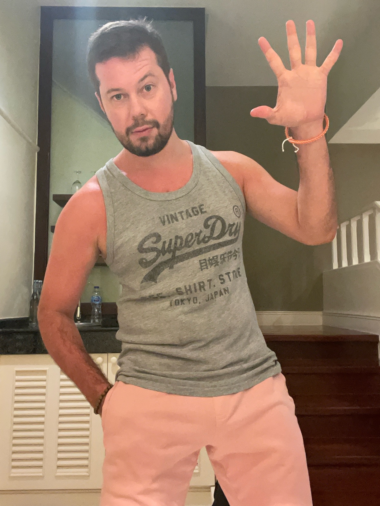 A man in a gray tank top and pink pants poses for a photo. He is holding up his hand in a greeting.