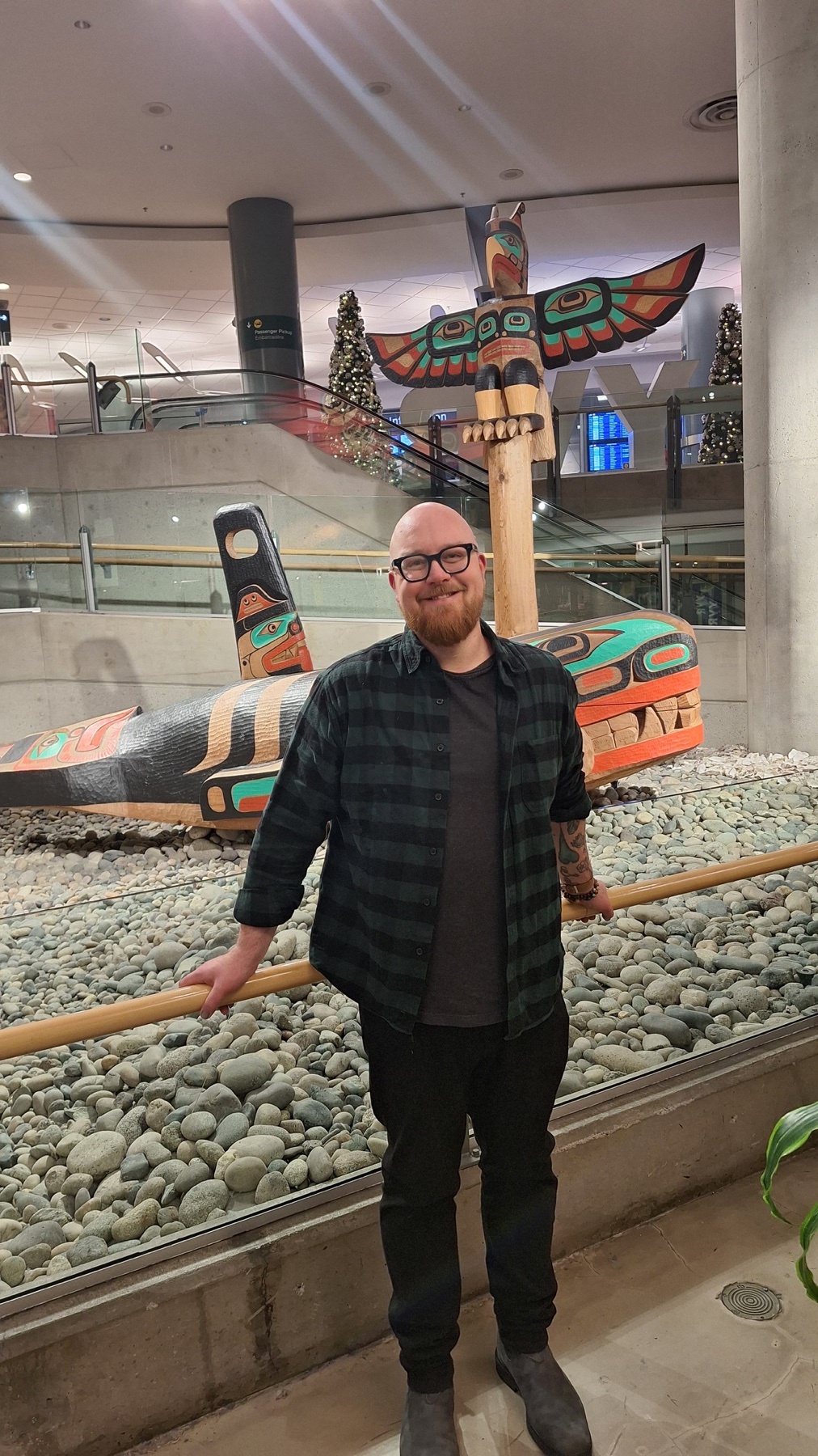 A man with glasses, a beard, and tattoos stands in front of a totem pole display. He is wearing a plaid shirt and dark pants.