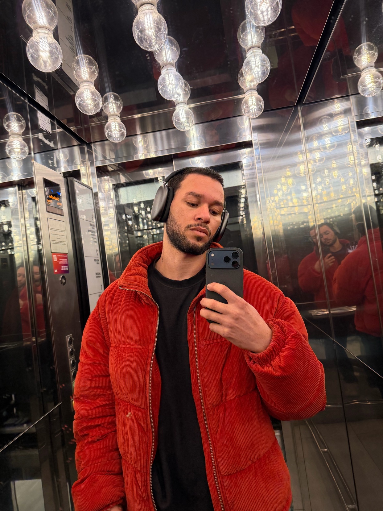 A man takes a selfie in a mirrored elevator, wearing a red jacket and headphones. The lighting is provided by multiple light bulbs.