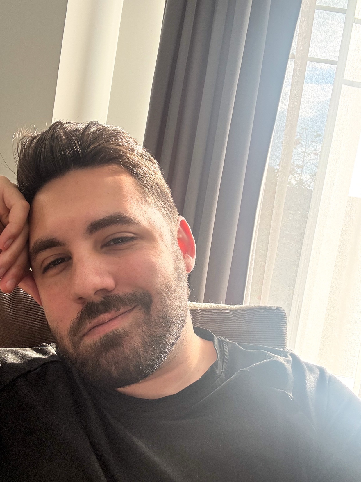 A man with a beard and dark hair smiles for a selfie, resting his head on his hand. He is wearing a black t-shirt and is indoors near a window with curtains.
