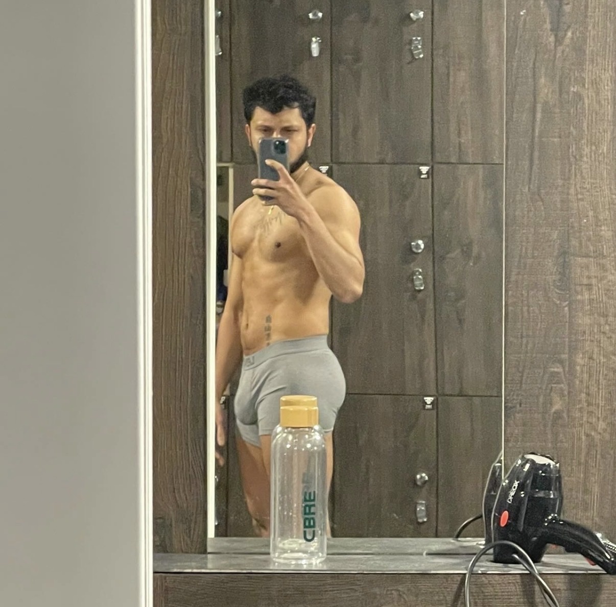 A man in grey underwear takes a mirror selfie in a gym locker room. He is muscular and has tattoos on his torso. A water bottle and hair dryer are visible in the reflection.