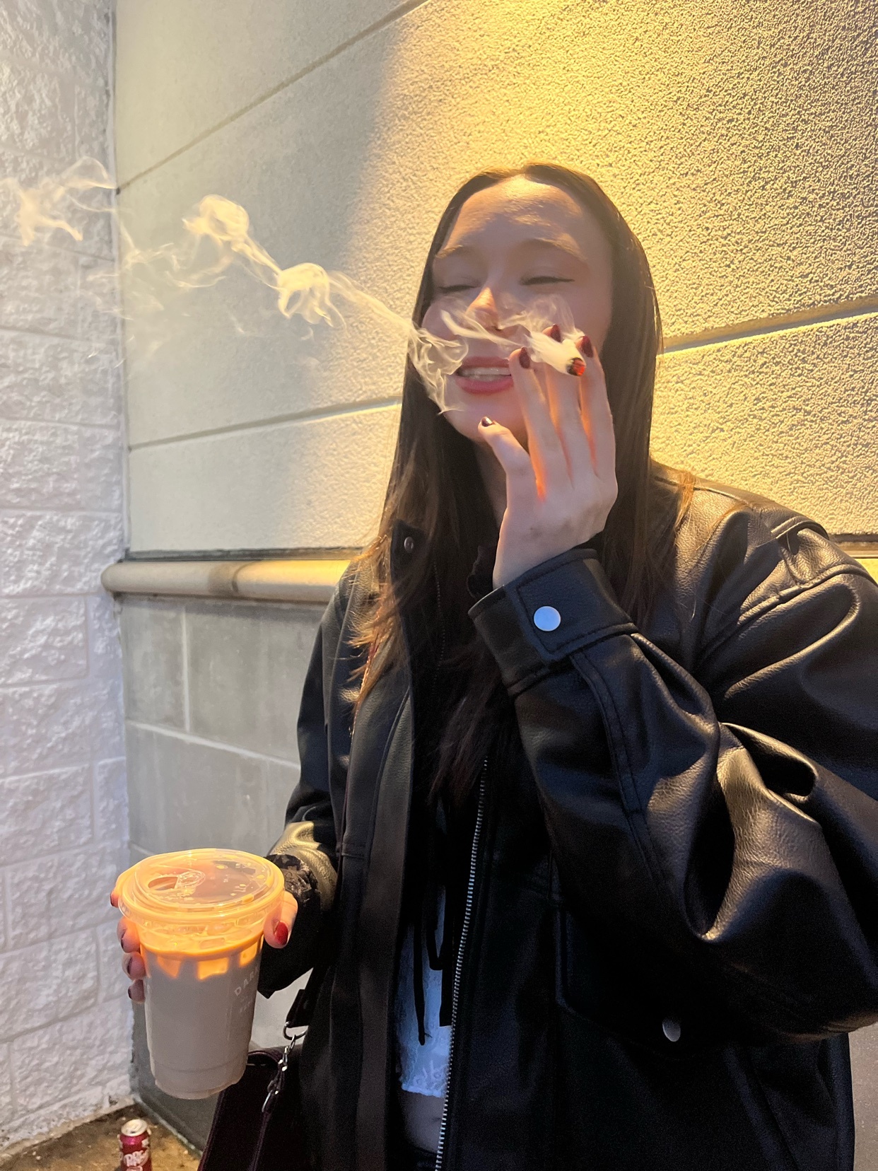 A young woman with long dark hair exhales smoke from a cigarette while holding a coffee cup. She is wearing a black leather jacket and is illuminated by warm light against a textured wall.
