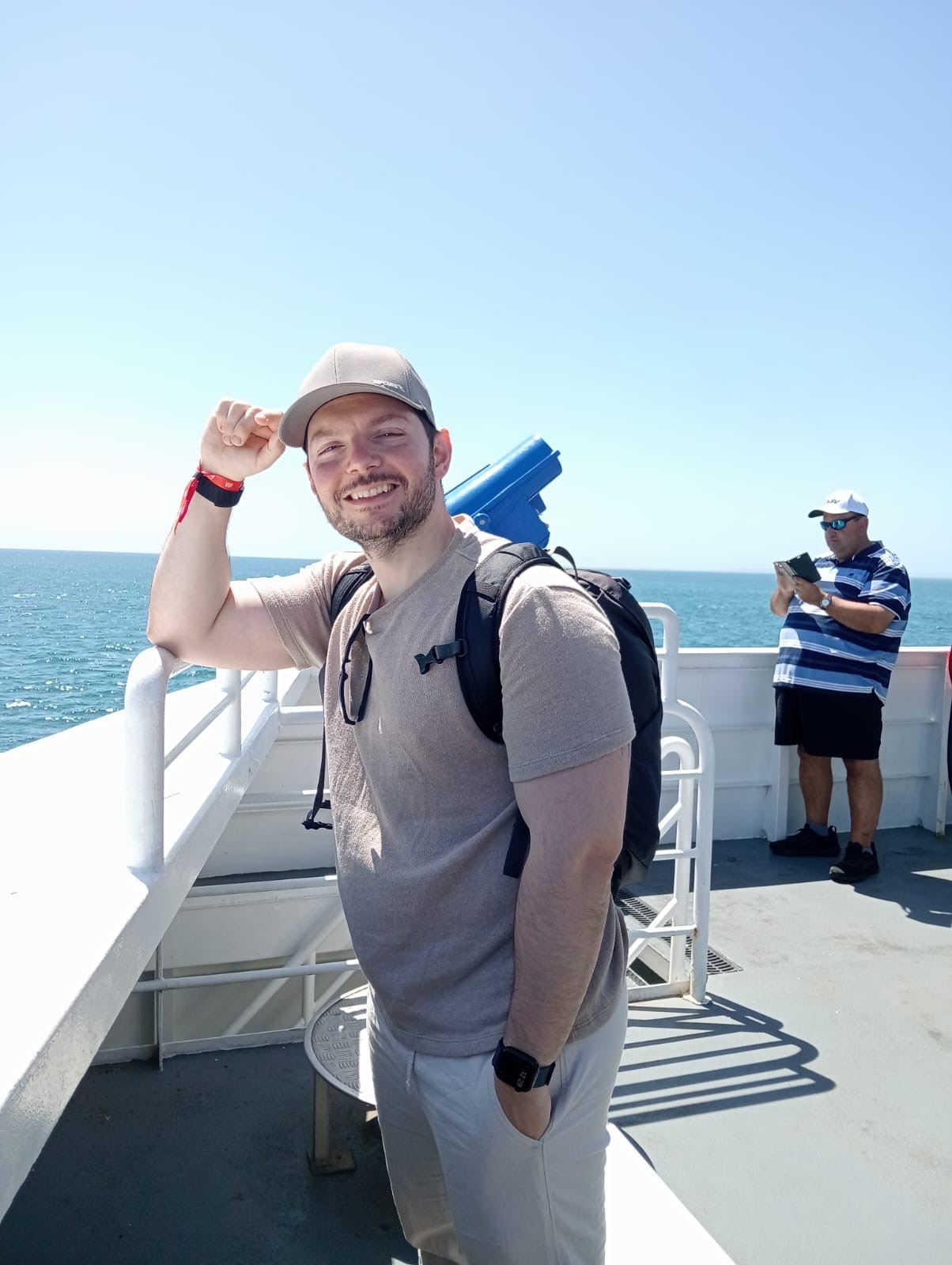 A man wearing a baseball cap and a t-shirt poses for a photo on the deck of a ferry with the ocean in the background.