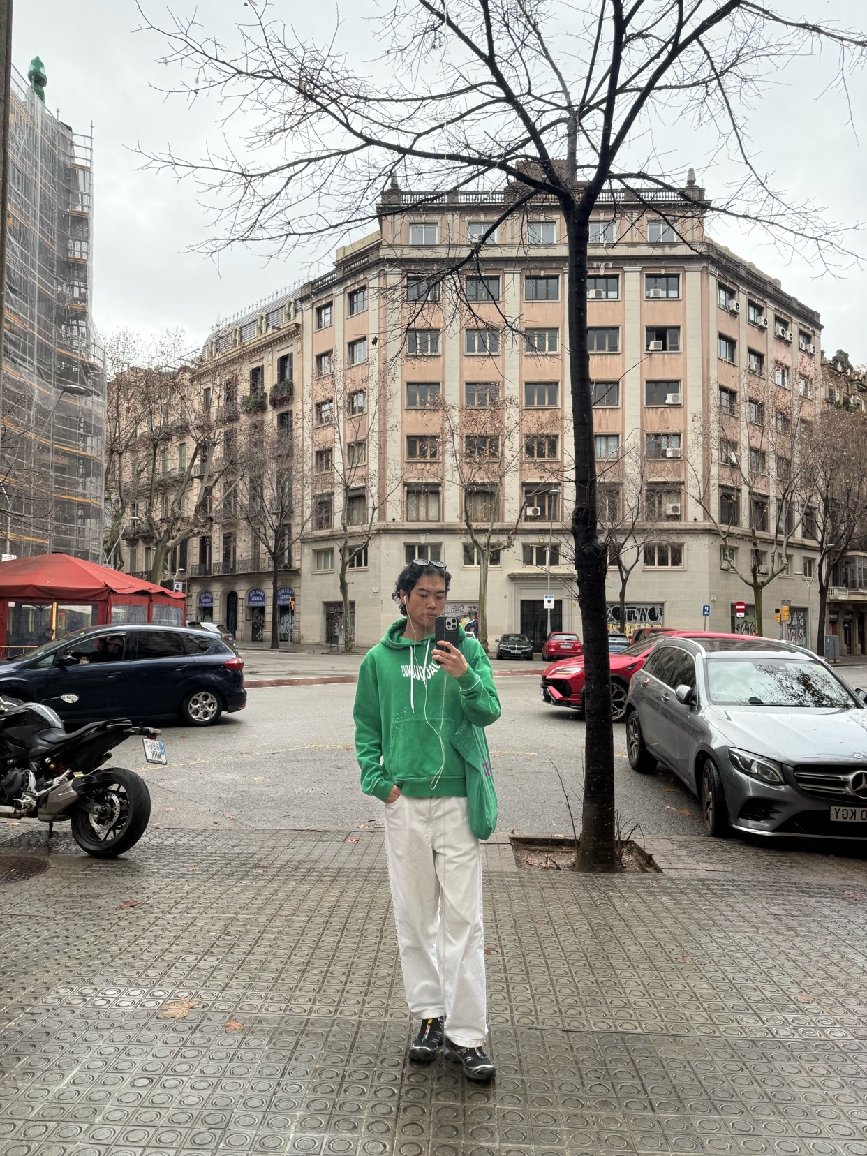 A man poses for a selfie on a city street. He wears a green hoodie, white pants, and sneakers.