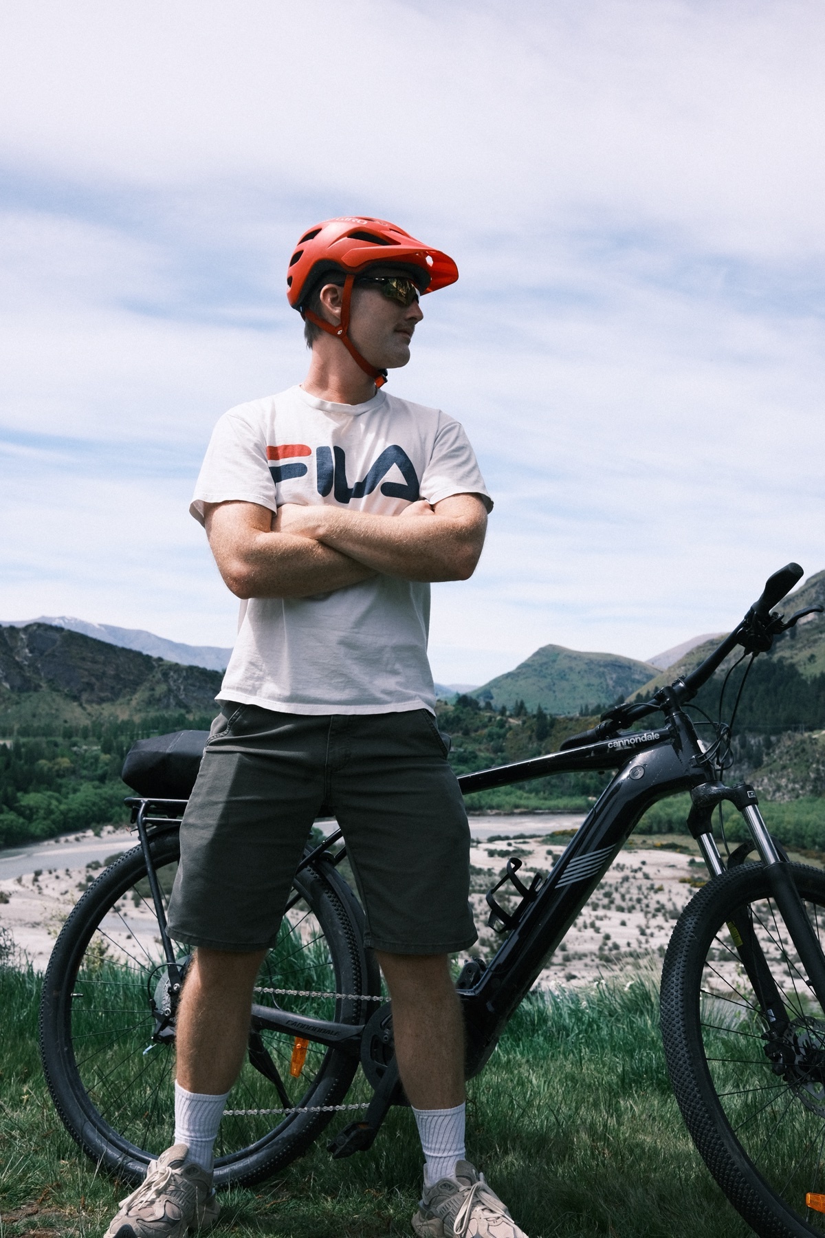 A man stands next to his mountain bike, wearing a helmet and sunglasses. He is posing with his arms crossed, with a scenic mountain backdrop.