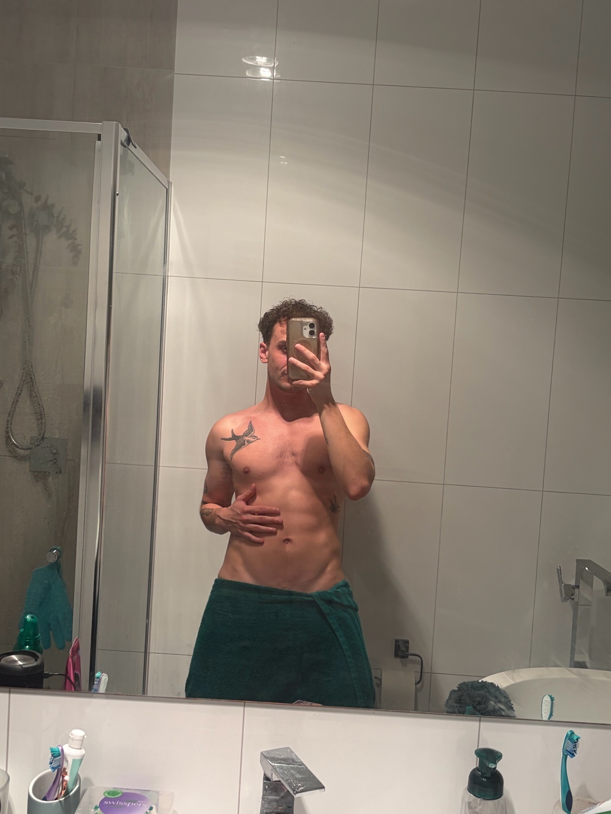 A man with a muscular physique and abs poses for a mirror selfie in a bathroom. He is shirtless and wearing a towel around his waist, with a tattoo visible on his chest.