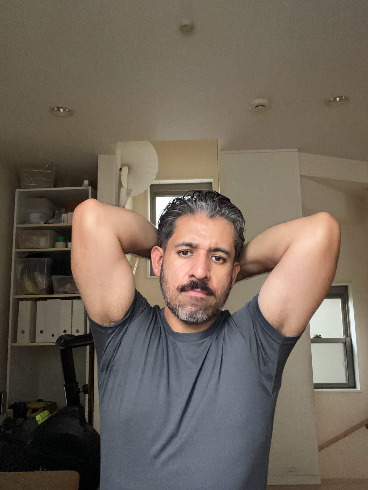 A man with a mustache and beard poses for a selfie indoors, with his arms behind his head.