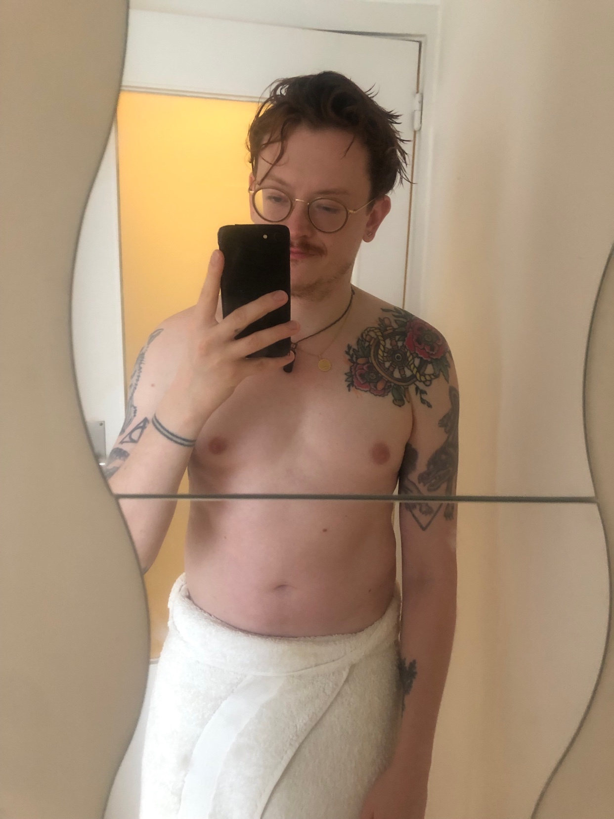 A man takes a mirror selfie, shirtless and wrapped in a towel. He has tattoos on his arm and shoulder.