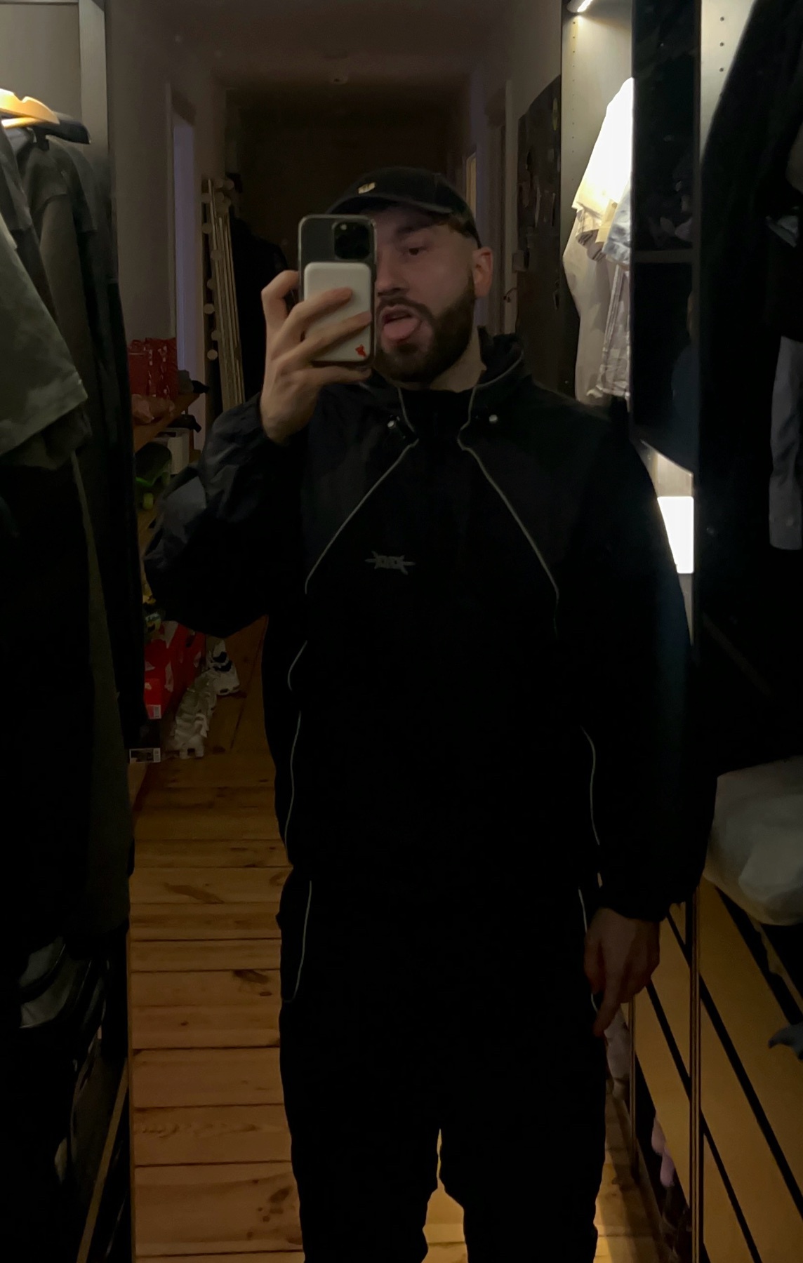 A man takes a mirror selfie in a closet, wearing a black jacket and cap. The photo showcases a modern streetwear style.