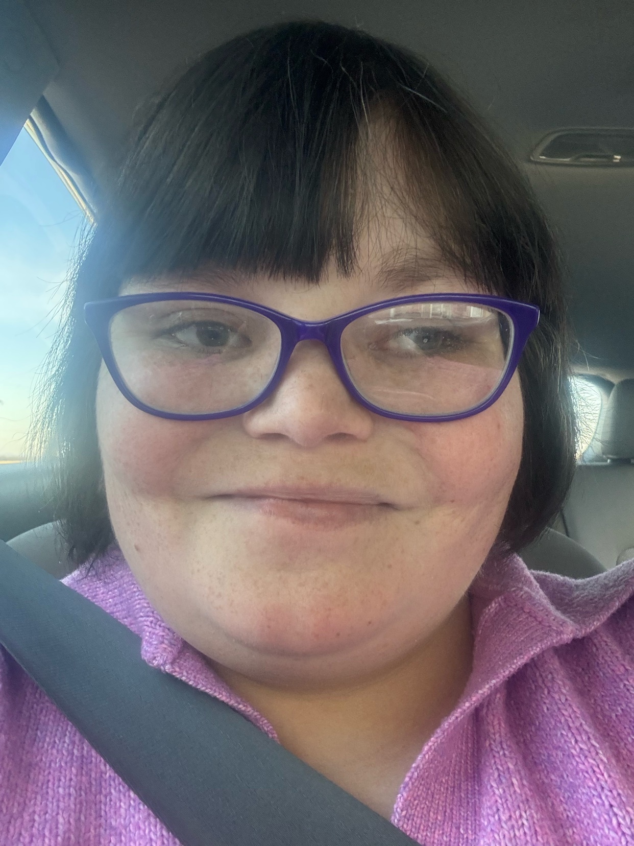 A woman with glasses and short dark hair takes a selfie in a car. She is looking directly at the camera.