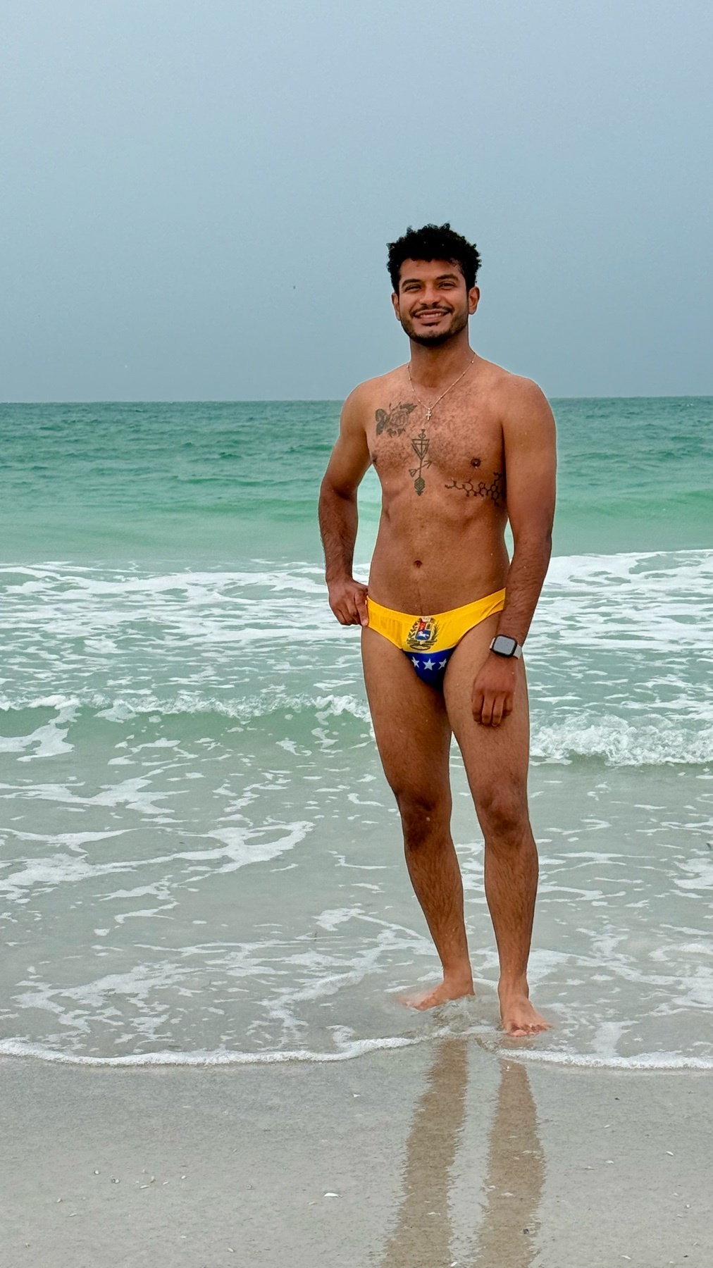A man stands in the ocean wearing a yellow speedo with the Venezuelan flag on it. He has tattoos on his chest and a watch on his left wrist.