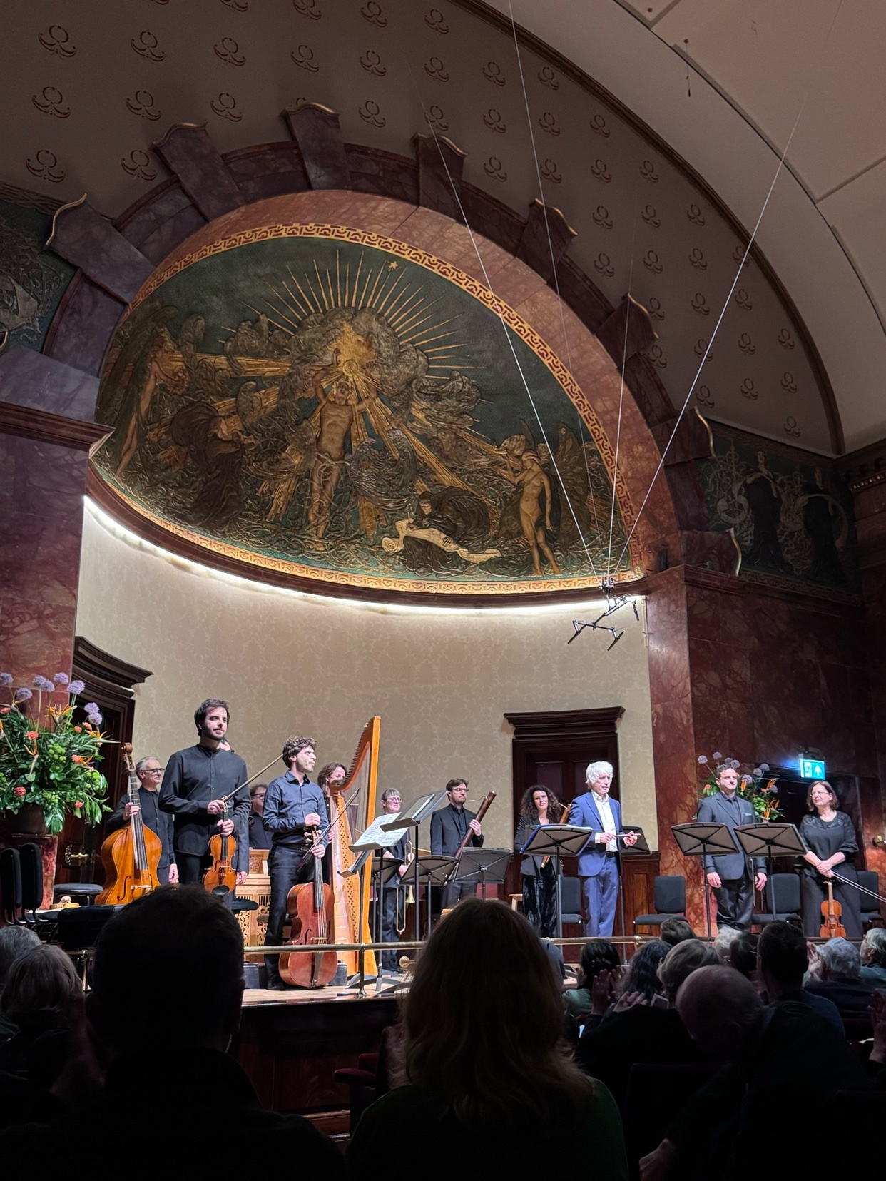 A classical music concert is taking place on a stage with a large mosaic above. The audience is visible in the foreground.
