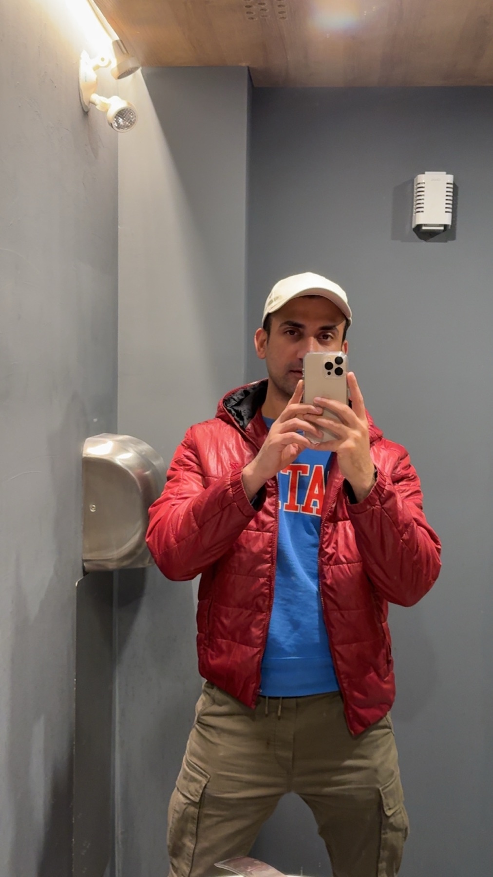A man takes a selfie in a public restroom, wearing a red puffy jacket over a blue t-shirt and a white baseball cap.