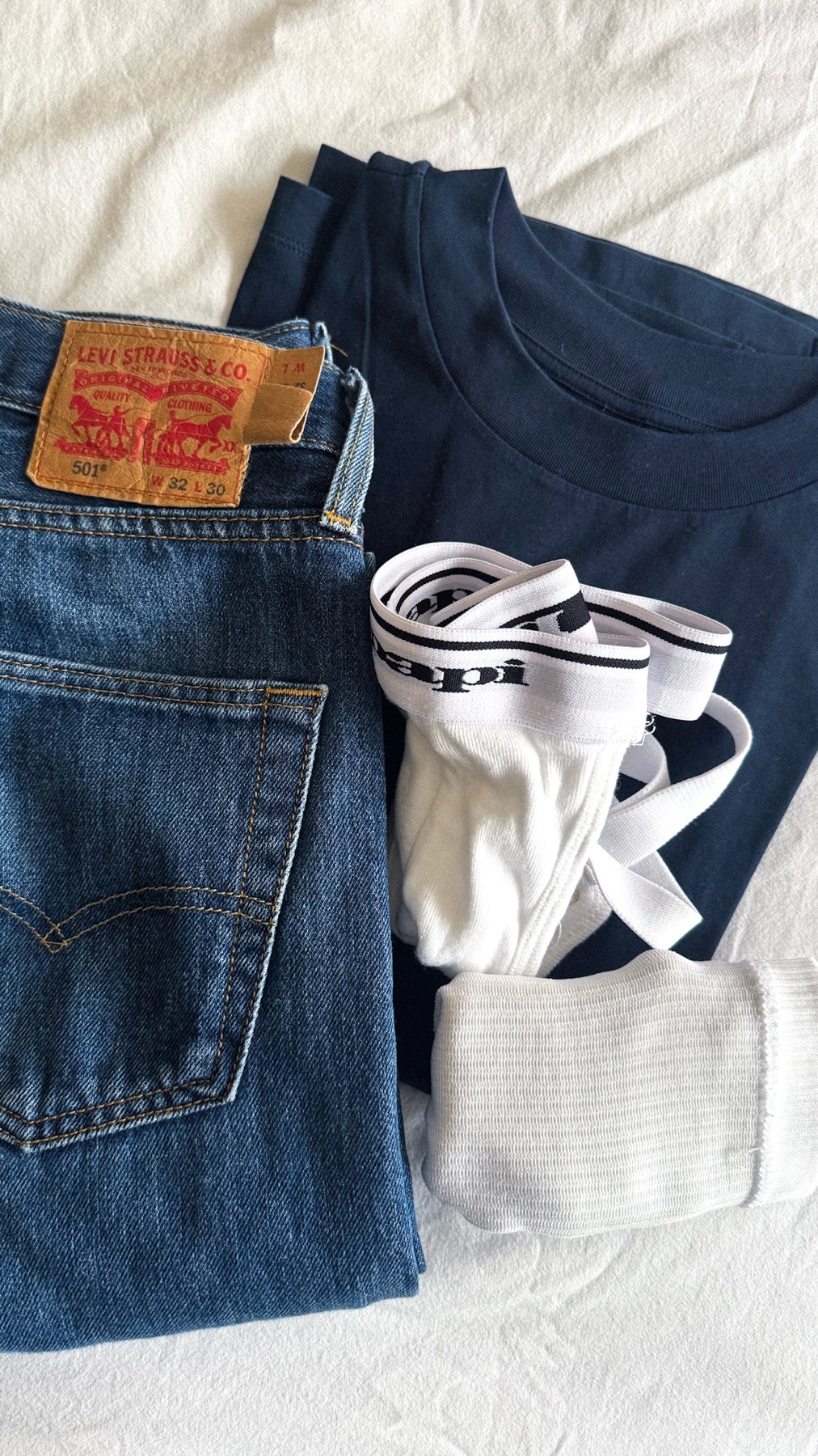 A flat lay of a men's outfit, including a pair of blue jeans, a navy blue t-shirt, and white underwear. The items are arranged on a white surface.