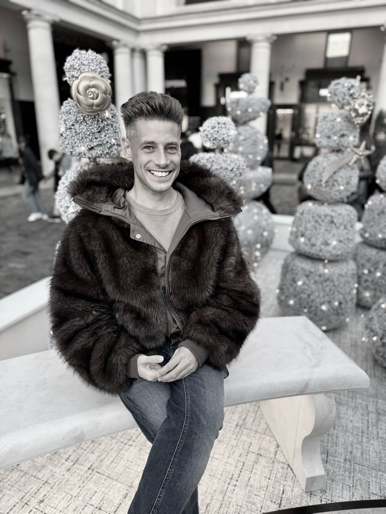 A man smiles while posing in a fur coat. He is sitting on a bench in front of some decorative holiday displays.