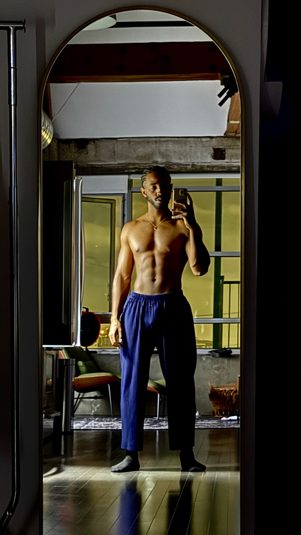 A shirtless man with a muscular physique poses for a mirror selfie indoors, wearing blue pants and socks.