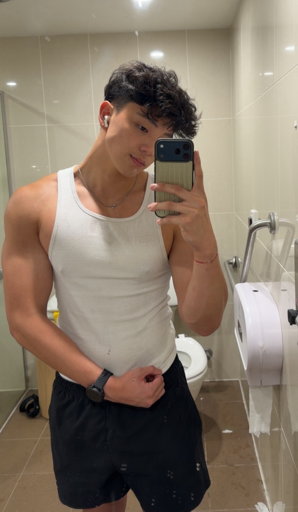 A man takes a mirror selfie in a bathroom, flexing his arm. He is shirtless, wearing a tank top and black shorts.