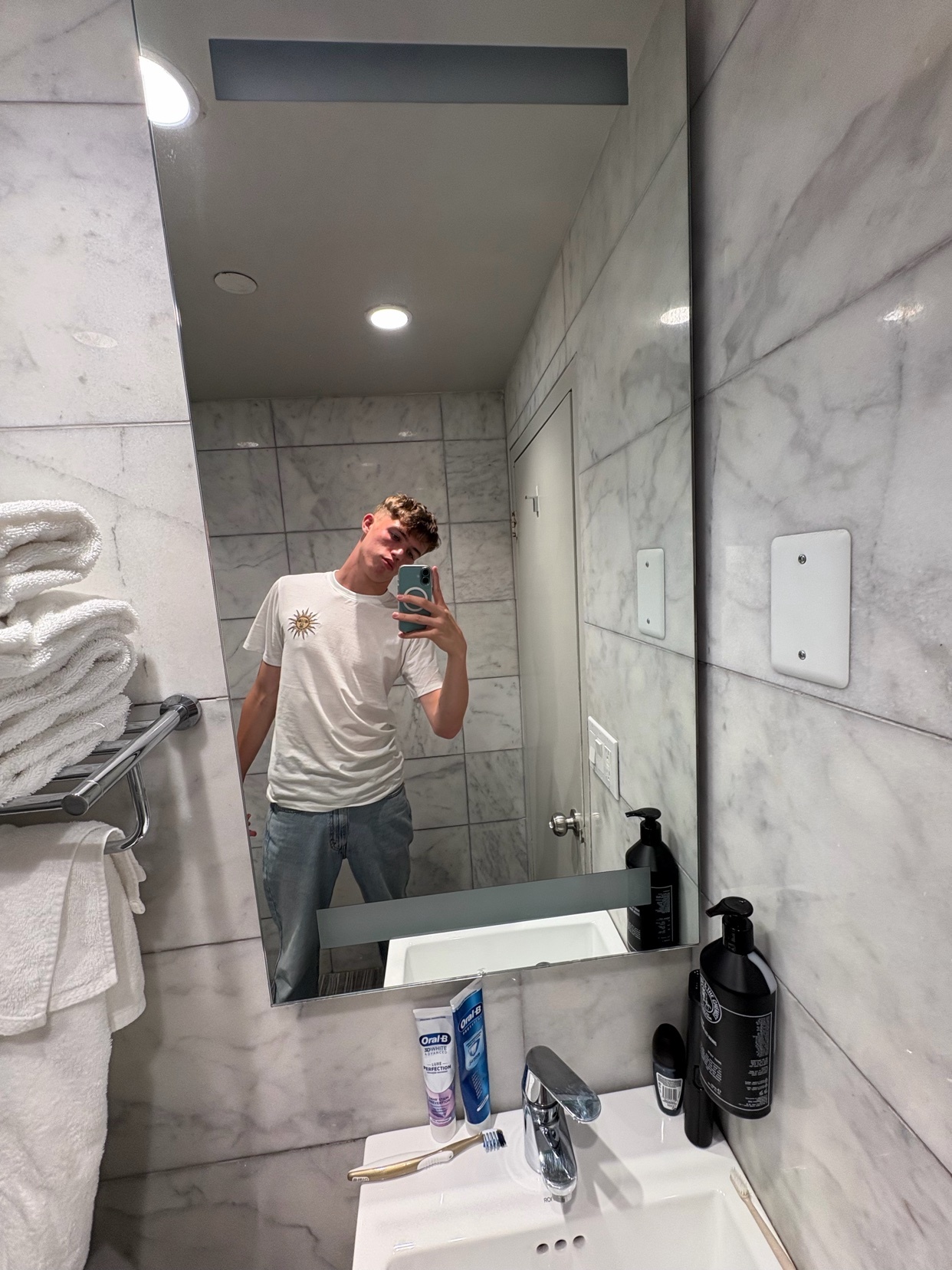 A young man takes a mirror selfie in a modern bathroom. He is wearing a white t-shirt and jeans, with blonde, short hair.